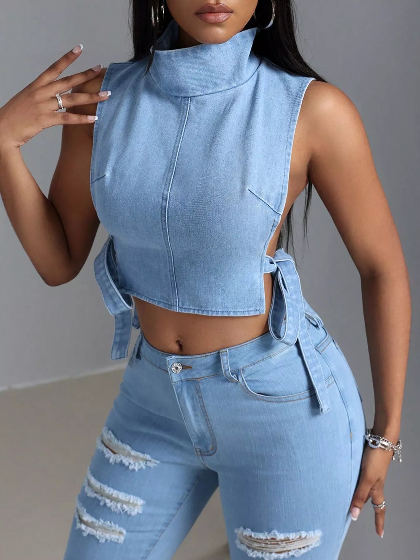 Woman wearing a light blue denim sleeveless top and high-waisted jeans against a gray background