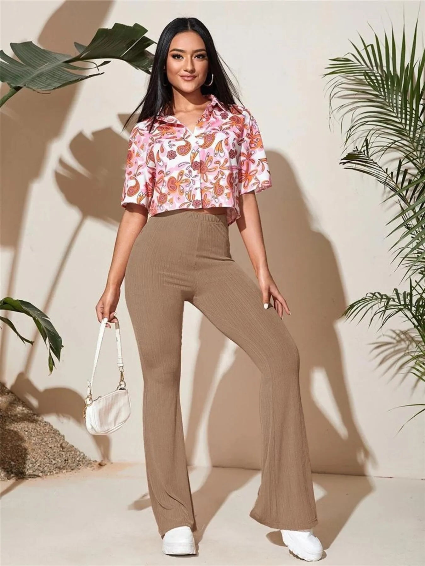 Woman wearing a floral blouse and brown pants standing against a neutral background with plant