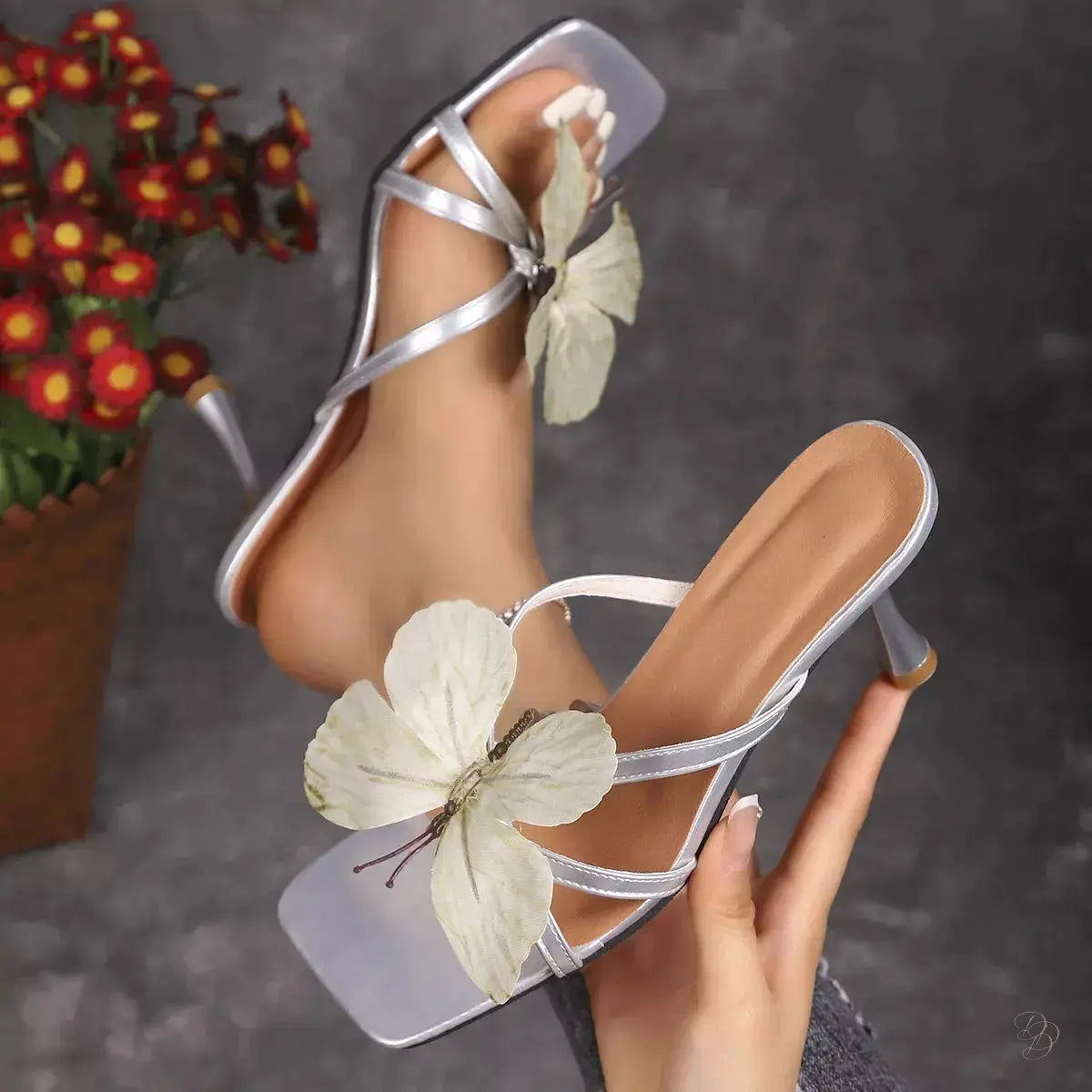 silver high-heeled sandals with floral decorations held by a hand against a dark background.