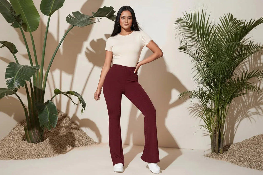 Woman wearing a floral top and burgundy pants with a white handbag, standing against a beige wall with plants.